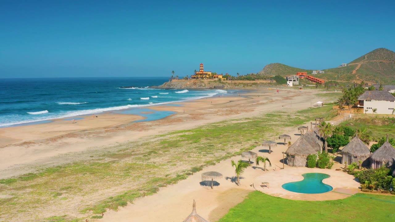 Mid-clip aerial frame over Cerritos with more foreground umbrellas visible near the pool, the beach running diagonally toward the distant coastal buildings, and no visible people.