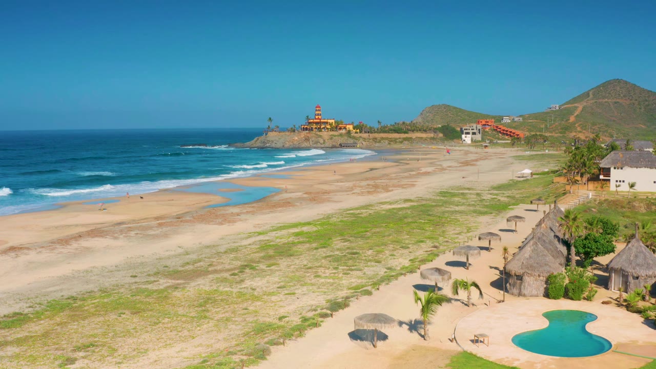 Cerritos beach aerial frame with the surf line slightly farther along the sand, shallow blue water channels visible near the center, huts and palms on the right, and no visible people.
