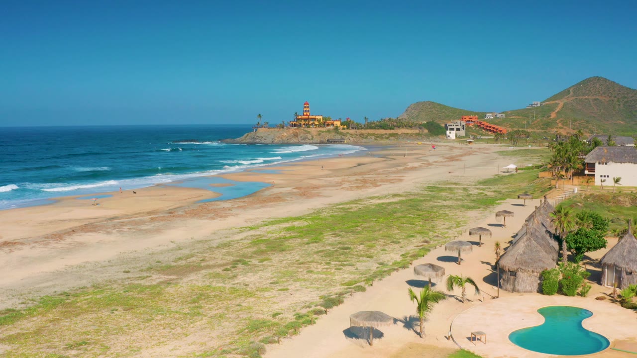 Aerial frame over Cerritos beach showing a long empty sand stretch, white wave lines on the left, a row of palapa-style umbrellas at right, hillside buildings beyond, and no visible people.