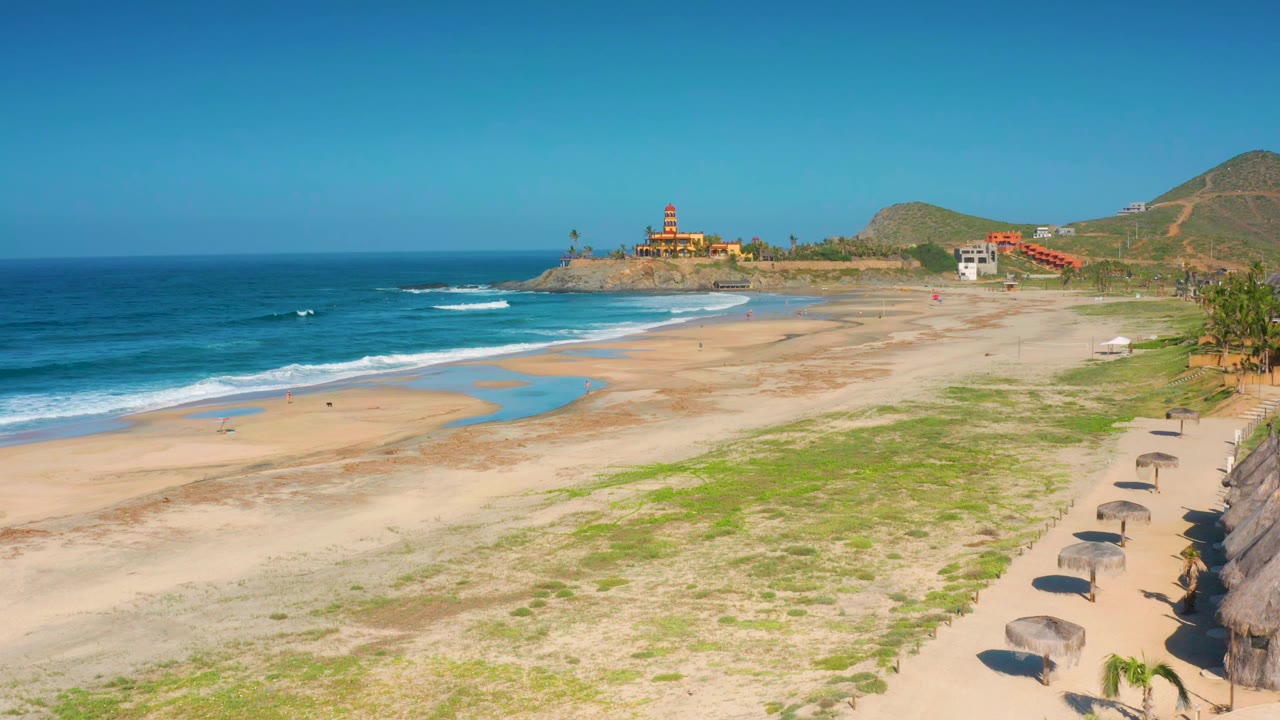 Cerritos beach remains wide in frame as small waves break near the shore, pale sand and green vegetation fill the midground, umbrellas line the right side, and no visible people appear.