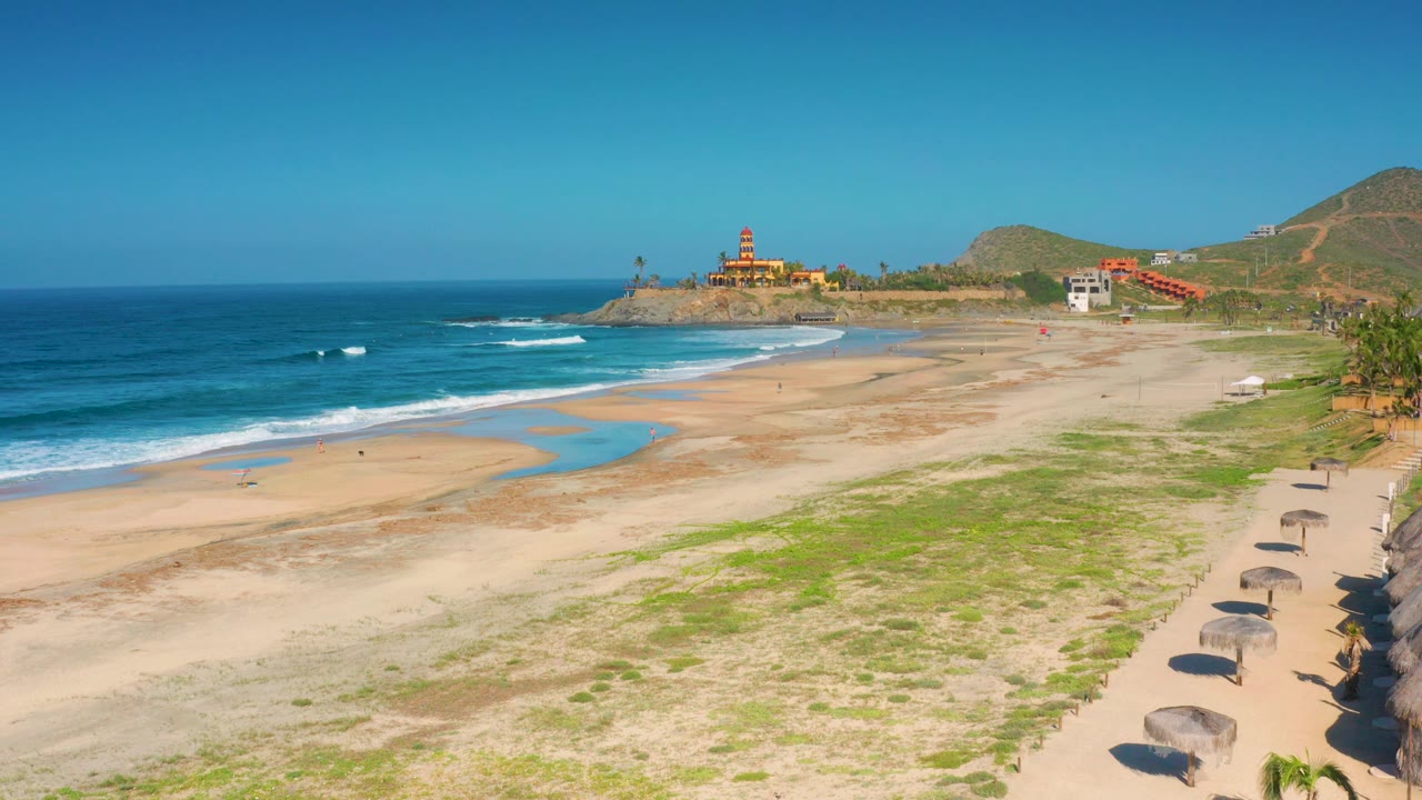 A high view over Cerritos, Baja California Sur, Mexico, shows turquoise water meeting broad sand, a curved wet area near the surf, umbrellas at the lower right, hillside buildings in the distance, and no visible people.