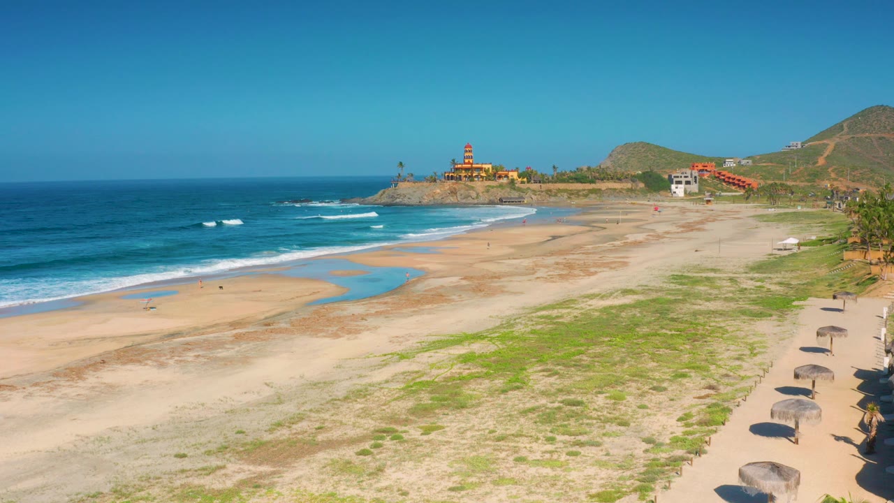 Mid-clip frame over Cerritos beach with mild waves rolling in from the left, long pale sand across the center, scattered green growth, arranged shade umbrellas along the right edge, and no visible people.