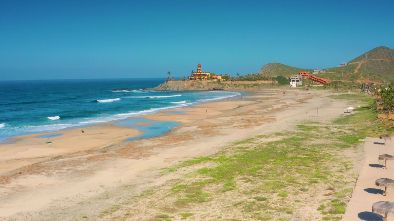 Opening view over Cerritos, Baja California Sur with blue surf at left, broad sand and shallow water channels below, hills beyond, a fort-like structure on the point, and no visible people.