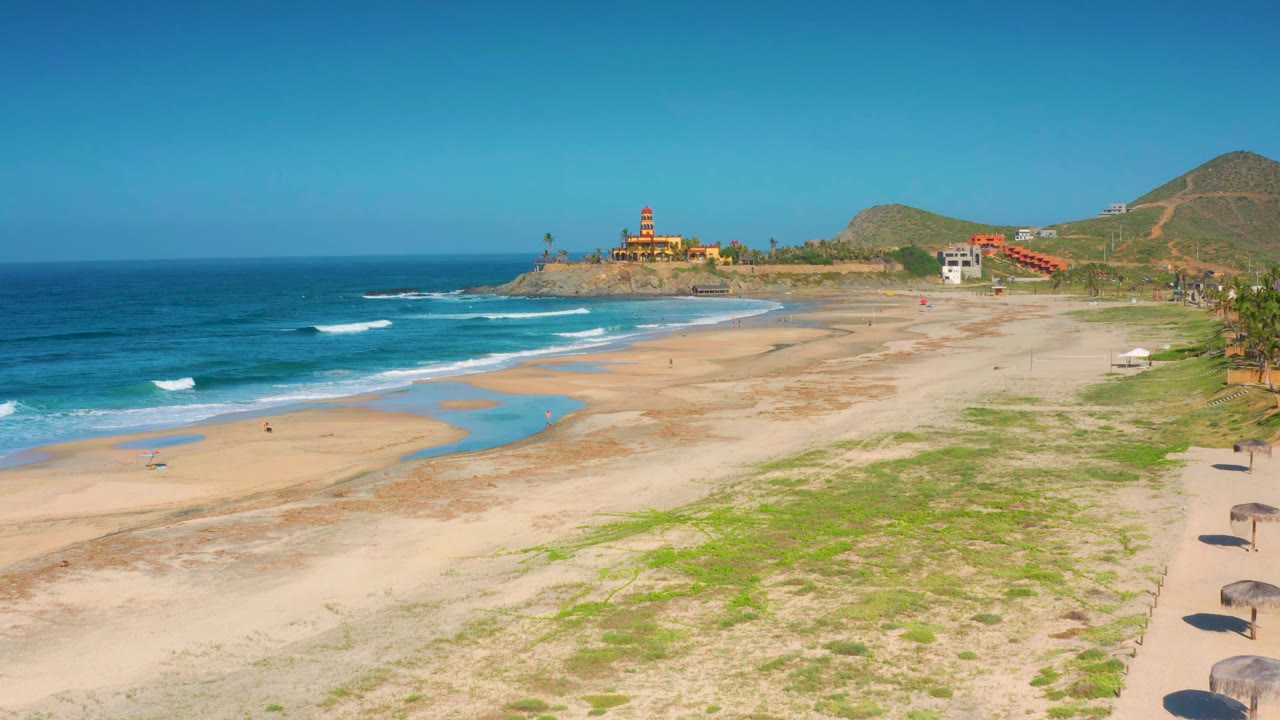 Cerritos coastline frame with small white wave lines moving toward open sand, green scrub in the foreground, hillside buildings, the fort-like structure near the horizon, and no visible people.