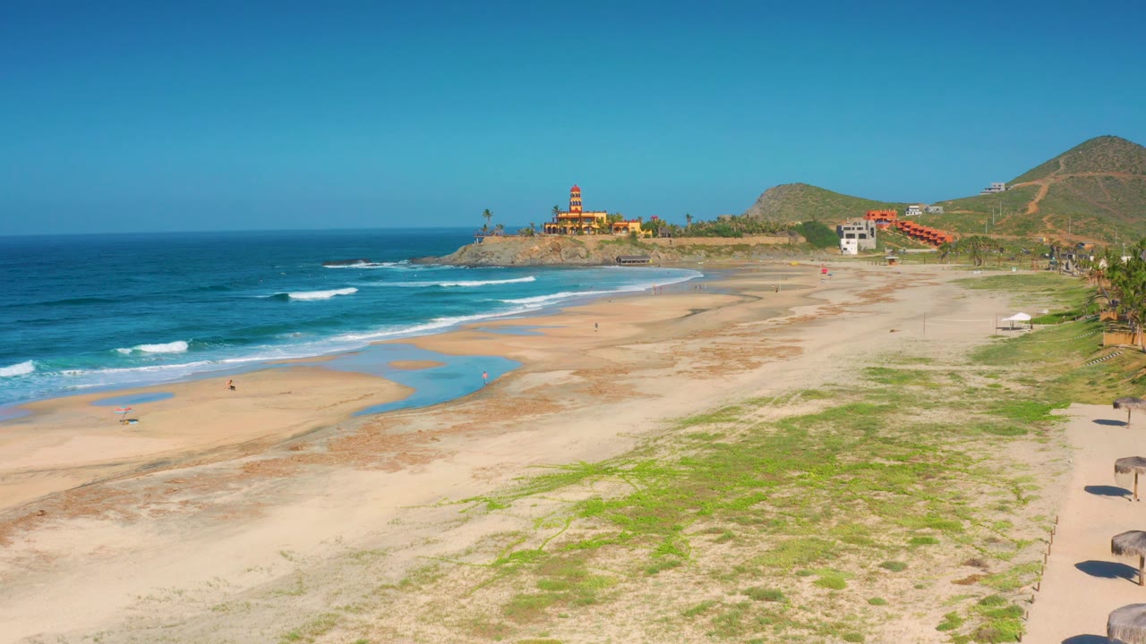 Wide Cerritos beach panorama showing shallow blue water across tan sand, surf on the left, a red-roofed fort-like structure on the point, hillside terrain behind it, and no visible people.