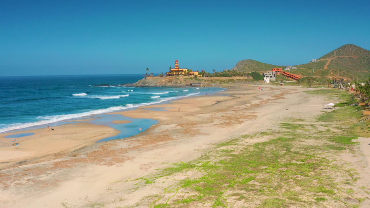 Opening frame of Cerritos, Baja California Sur with blue surf at left, pale sand and shallow pools across the beach, low hills behind coastal buildings, and no visible people identifiable.