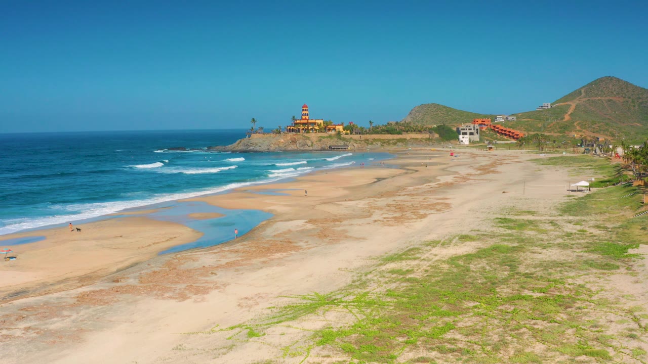 Elevated coastal view with beach pools, surf, vegetation, hills, and scattered buildings.