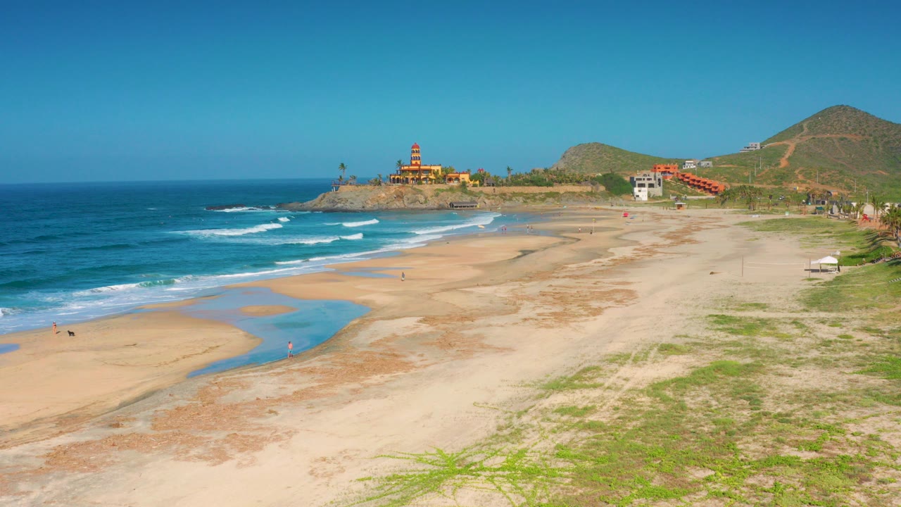 Opening frame of Cerritos, Baja California Sur beach with blue surf at left, pale sand in front, a red domed building on the rocky point, hills behind it, and no identifiable people visible.