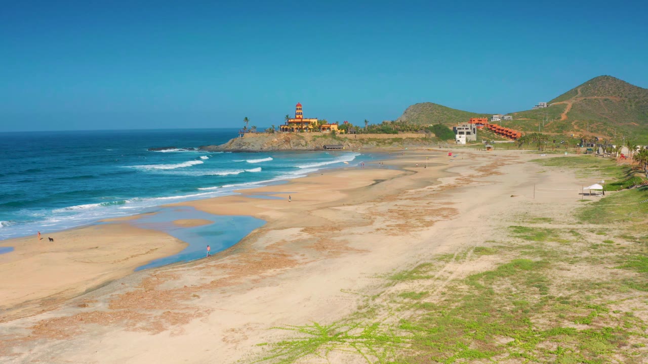 Sandy coastline with surf, tidal water, hills, and red domed building.