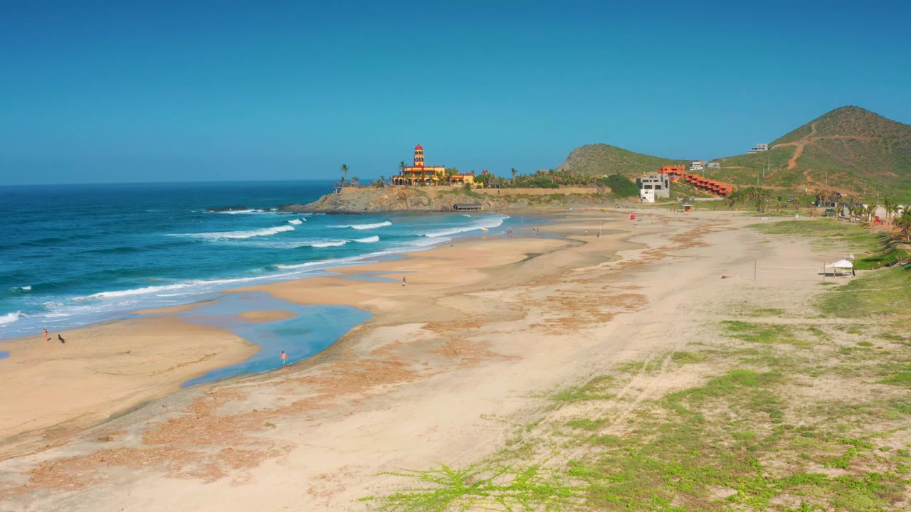Cerritos beach frame with white wave bands farther along the left shoreline, the red domed building on the point, smaller coastal buildings to the right, and no identifiable people visible.