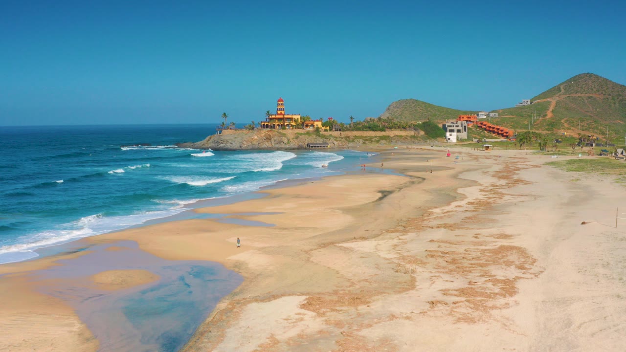 A wide aerial frame over Cerritos, Baja California Sur shows blue surf, wet sand pools, green hills, and a distant lighthouse on a rocky point with no visible people.