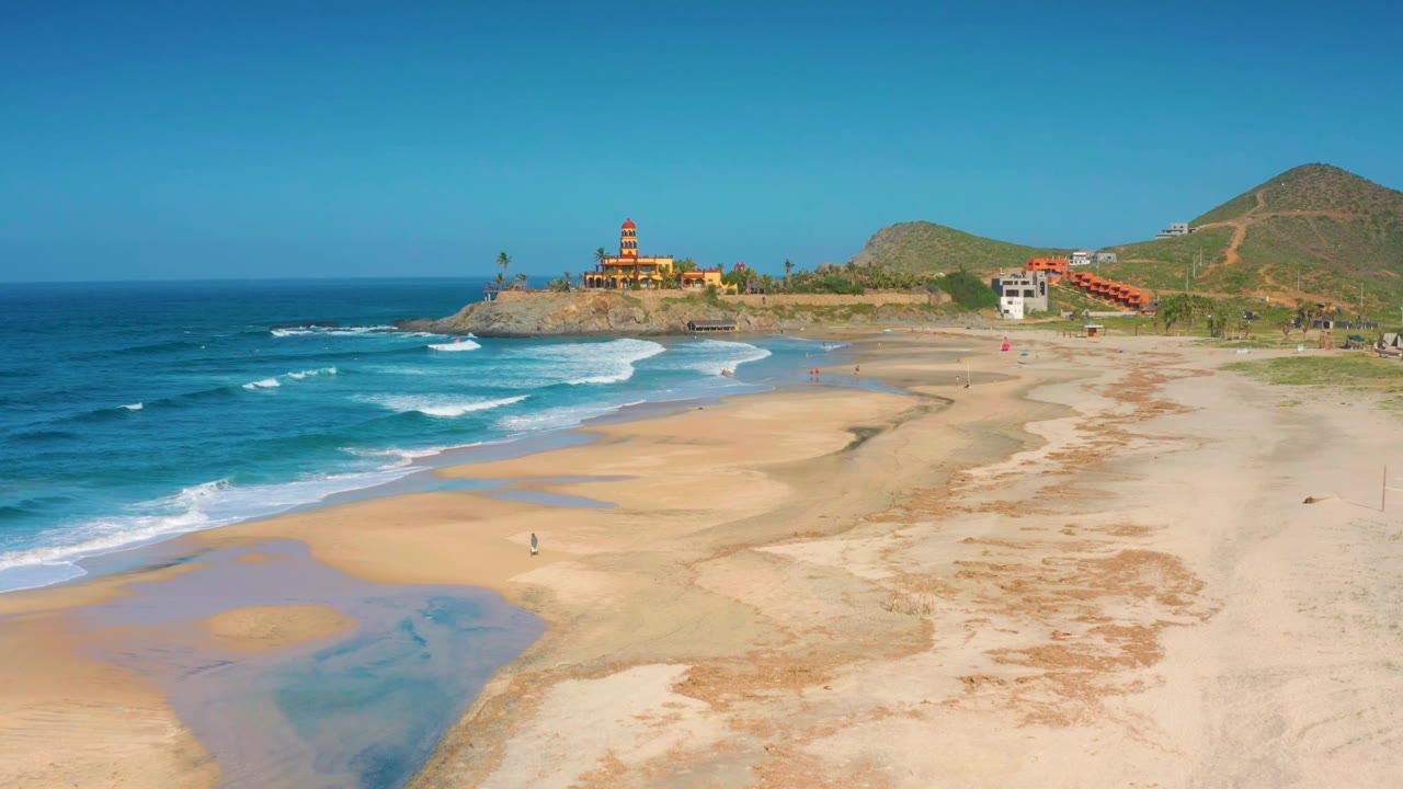 At 0:01, the Cerritos beach view holds the curving surf line, broad sand flats, shallow water patches, hillside buildings, and lighthouse under a clear sky with no visible people.
