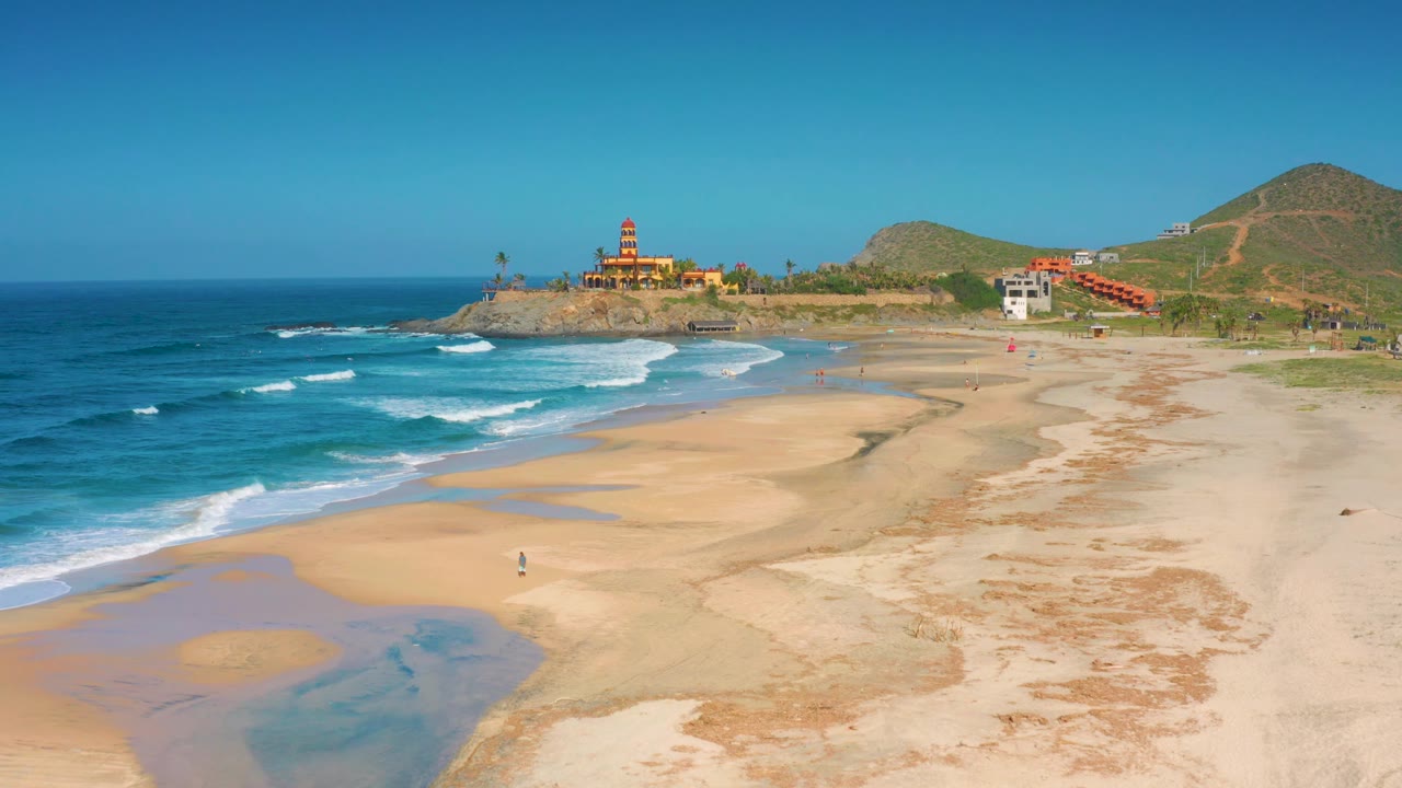 At 0:02, the aerial view shows more of the dry beach on the right, surf bands at left, sandy pools near the foreground, and the Cerritos lighthouse on the headland with no visible people.