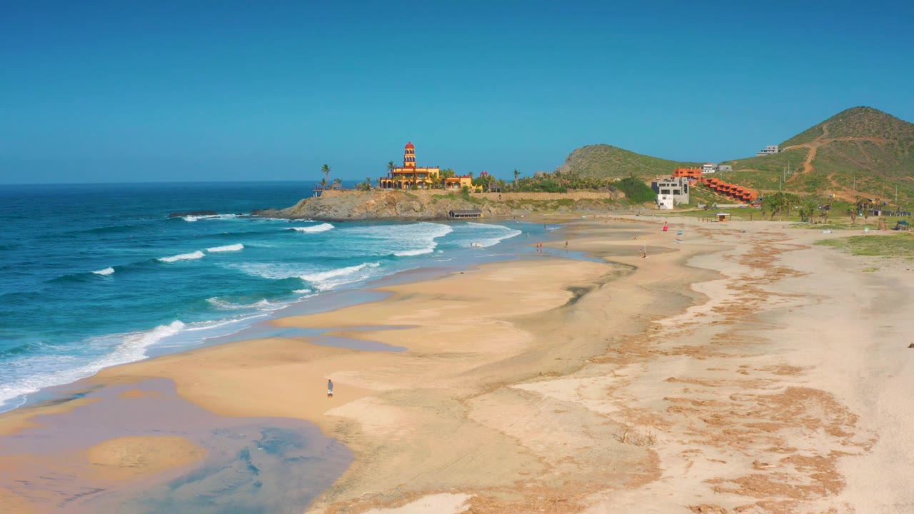 Elevated coastal view of waves, wet sand pools, beach, hills, and lighthouse.