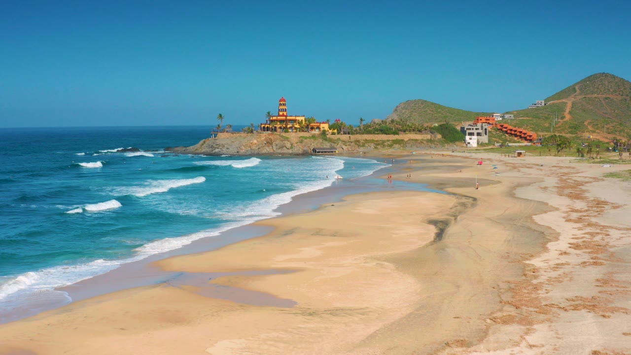 At 0:00, Cerritos beach appears from an elevated angle with tan sand in front, blue ocean to the left, shoreline buildings near the point, low hills beyond, and no visible people.