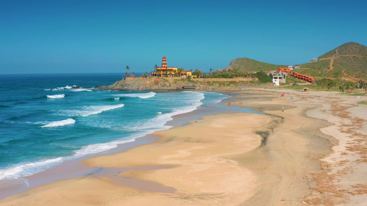 At 0:02, the Cerritos beach view shows more surf texture along the left edge, pale sand flats across the foreground, a cluster of coastal buildings near the hill, and no visible people.