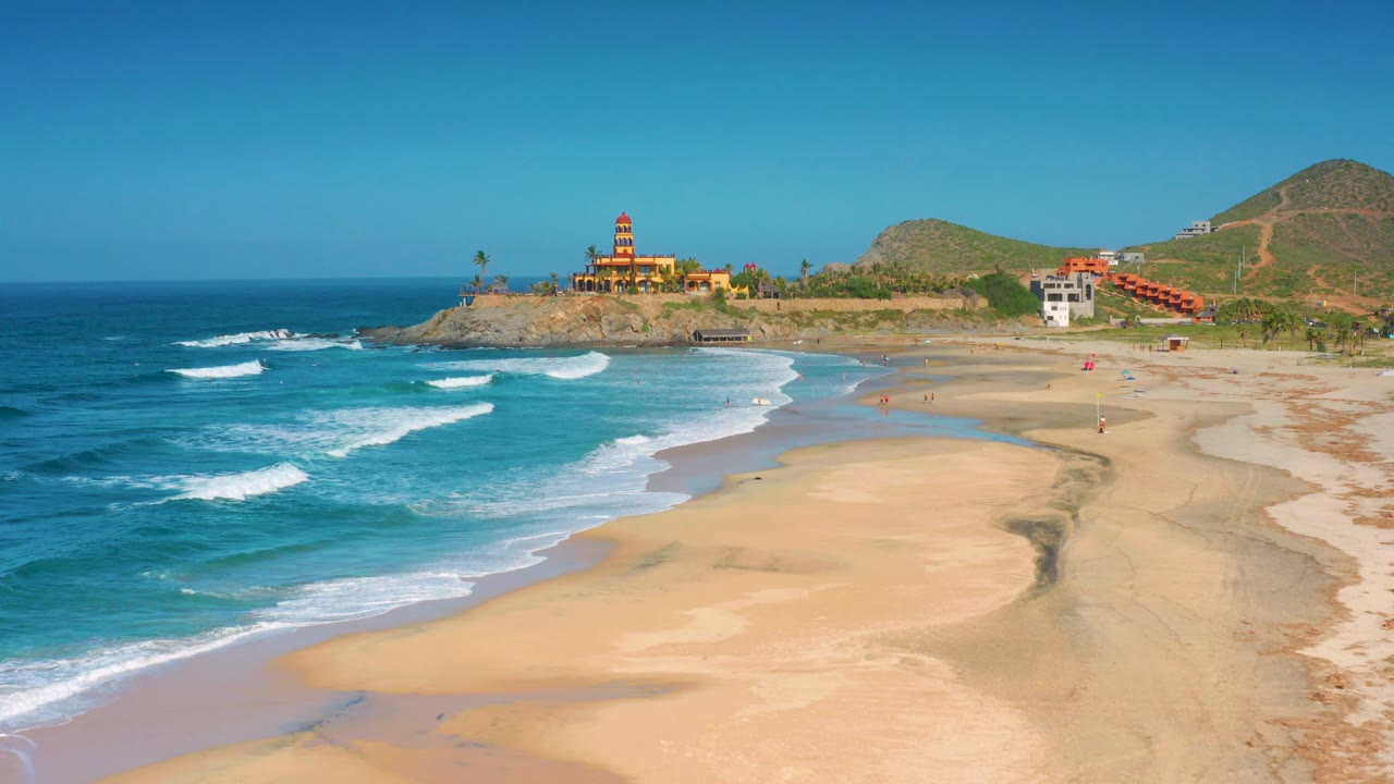 Elevated coastline view with waves, broad beach, rocky point, and hillside buildings.