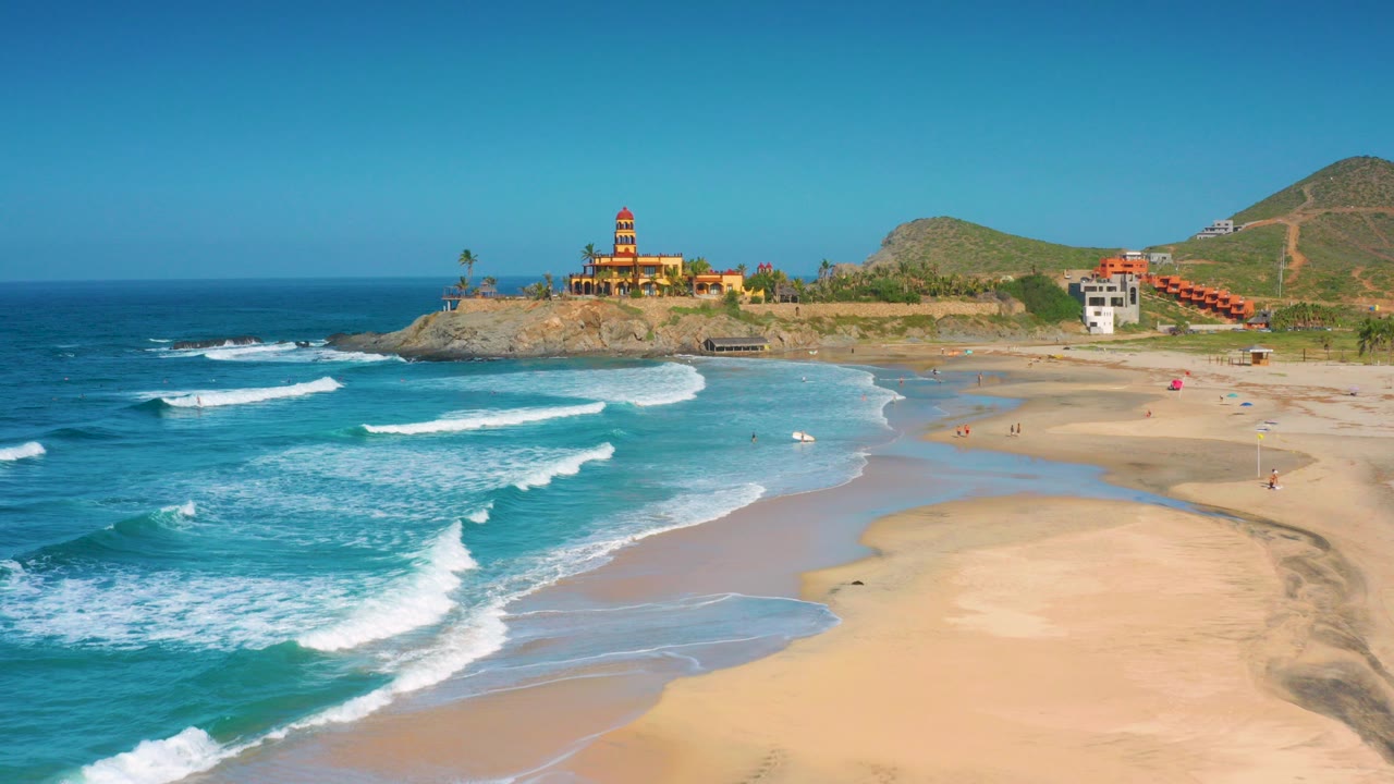 At 0:00, Cerritos beach appears from above with turquoise surf, broad sand, rocky shoreline, hillside buildings, and no identifiable people.