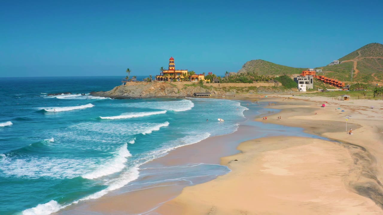 Wide aerial beach view with white waves curling toward wet sand and buildings on a rocky point.