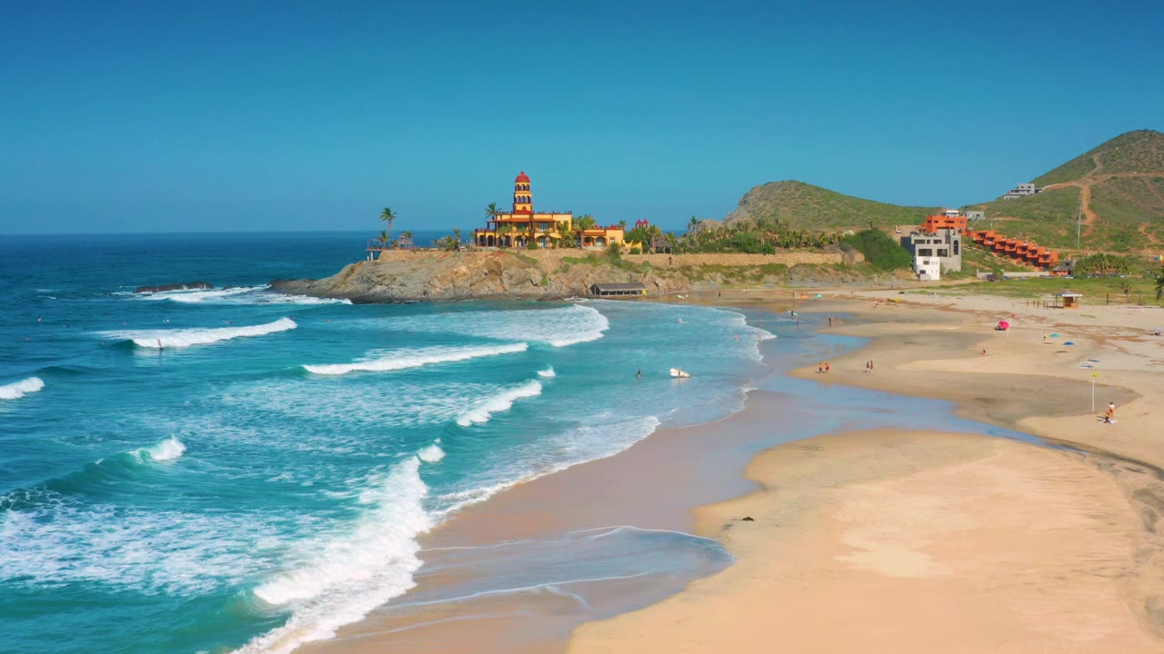 At 0:02, Cerritos beach is shown with diagonal surf lines, a shallow water edge, rocky headland buildings, hillside terrain, and no identifiable people.