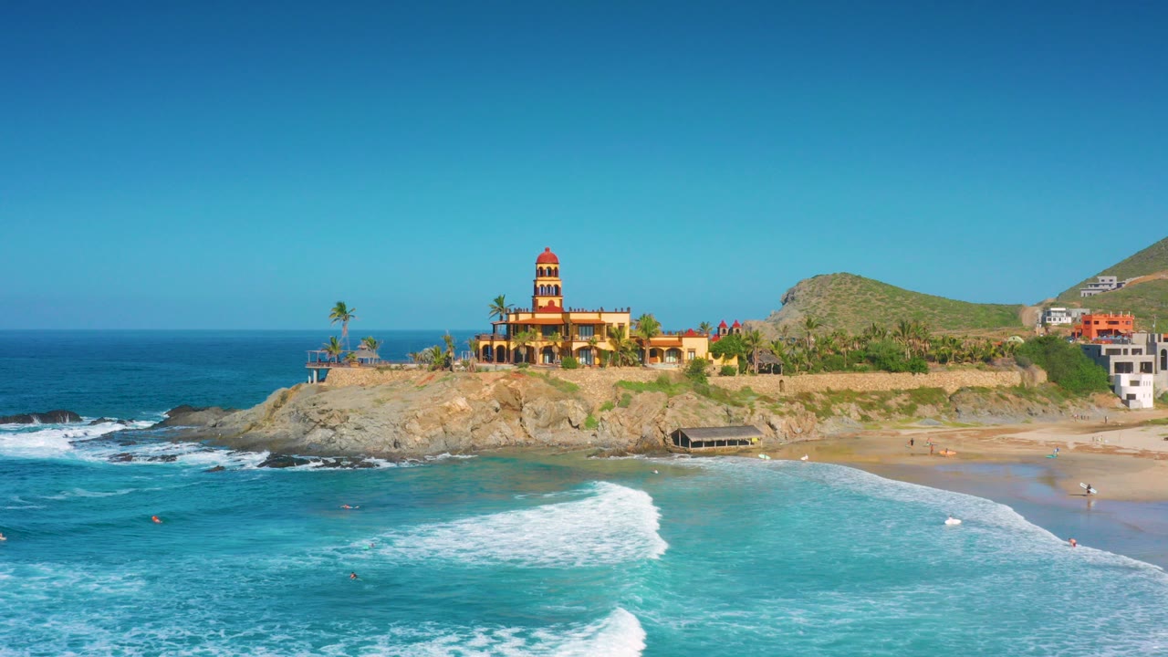 Opening aerial frame over Cerritos, Baja California Sur showing blue ocean, white surf, tan rocks, sandy beach, hillside buildings, and a tower on the coast with no visible people.