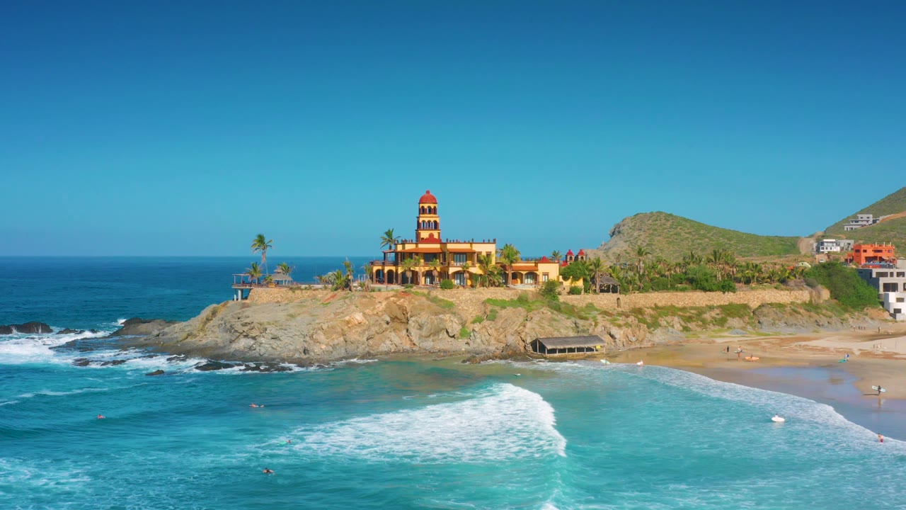 Cerritos aerial frame showing the wave foam shifted farther across the foreground water, with the tower, palms, rocky shore, sand, and hillside buildings in bright sun and no visible people.