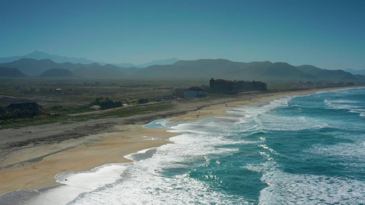 Aerial frame over Cerritos, Baja California Sur, showing white surf reaching a broad sandy beach, turquoise water to the right, hazy mountains inland, and no visible people.