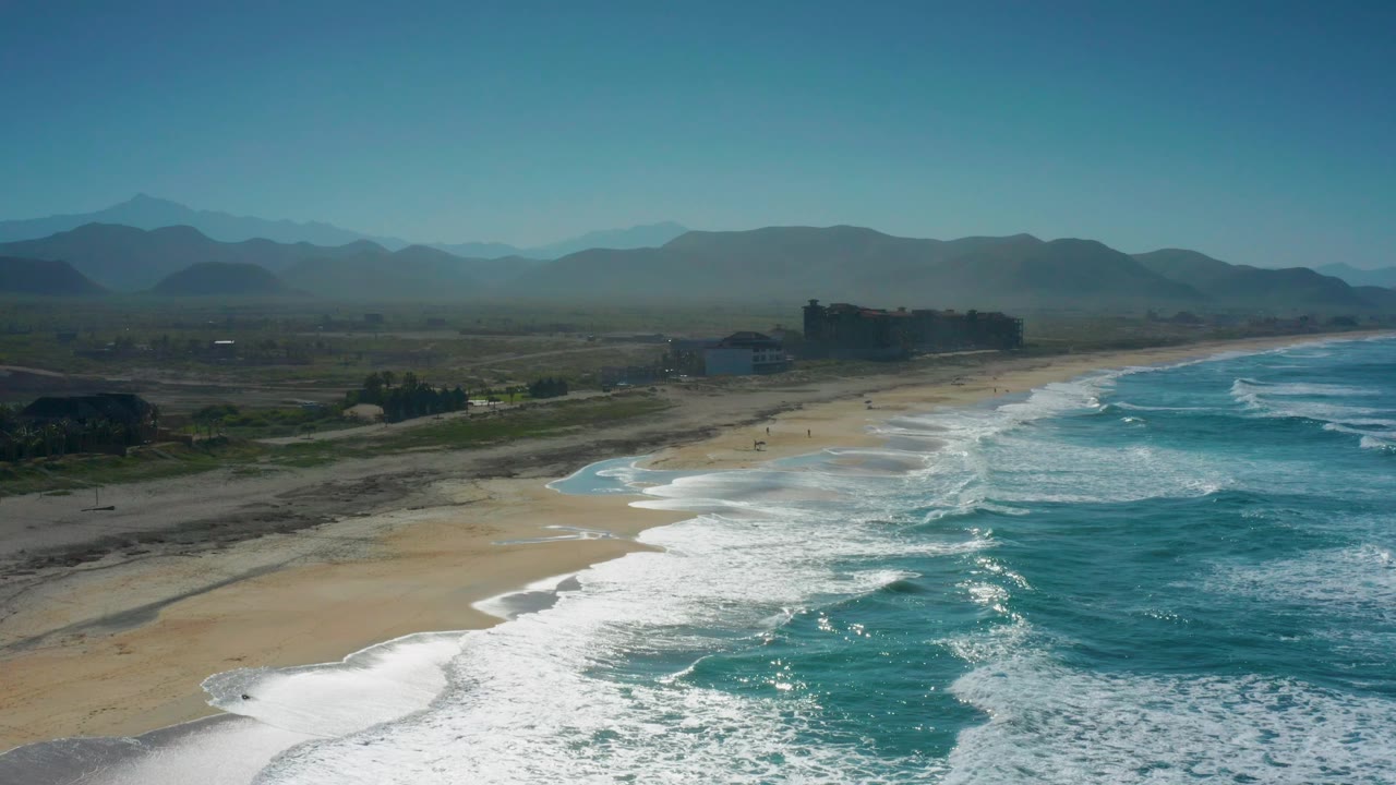 Cerritos, Baja California Sur coastline from above, with the surf line arcing across the lower frame, open sand through the center, hazy mountain ridges in back, and no visible people.