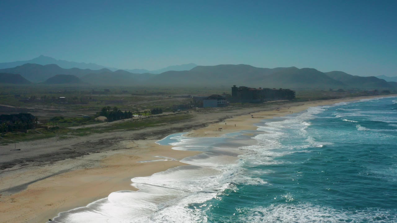 Aerial view over Cerritos, Baja California Sur, with white surf entering from the lower left, turquoise ocean on the right, sandy beach through the center, hazy mountains beyond, and no visible people.