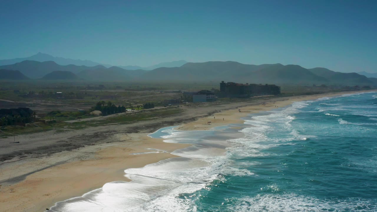 Cerritos shoreline seen from above as a broad white wave line spreads across the lower beach, with blue-green water to the right, low inland land to the left, distant structures, mountains, and no visible people.