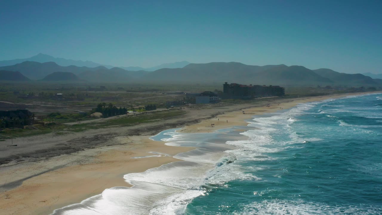 Aerial coastal view in Cerritos with brighter foam bands stepping up the beach, deeper turquoise water on the right, flat inland terrain and small distant buildings on the left, hazy mountain ridges, and no visible people.