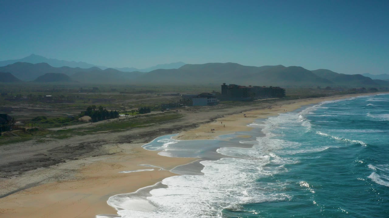 Aerial view of Cerritos, Baja California Sur showing turquoise waves breaking along a sandy beach, dark inland terrain, low shore buildings, hazy mountains, and no visible people.