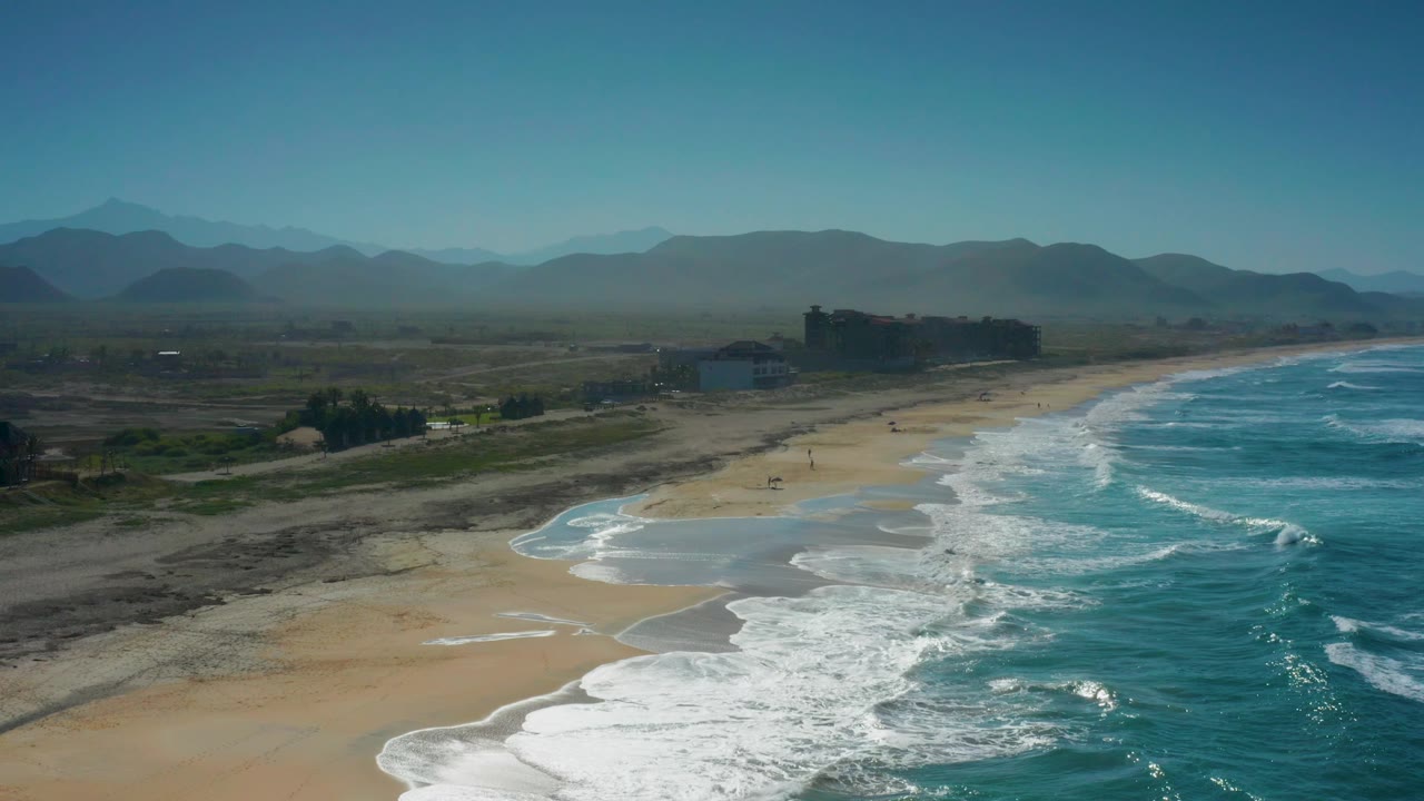 Cerritos coastline seen from above with a wide band of blue water and foam on the right, open sand at center, dark rocky terrain inland, shore buildings, mountains, and no visible people.