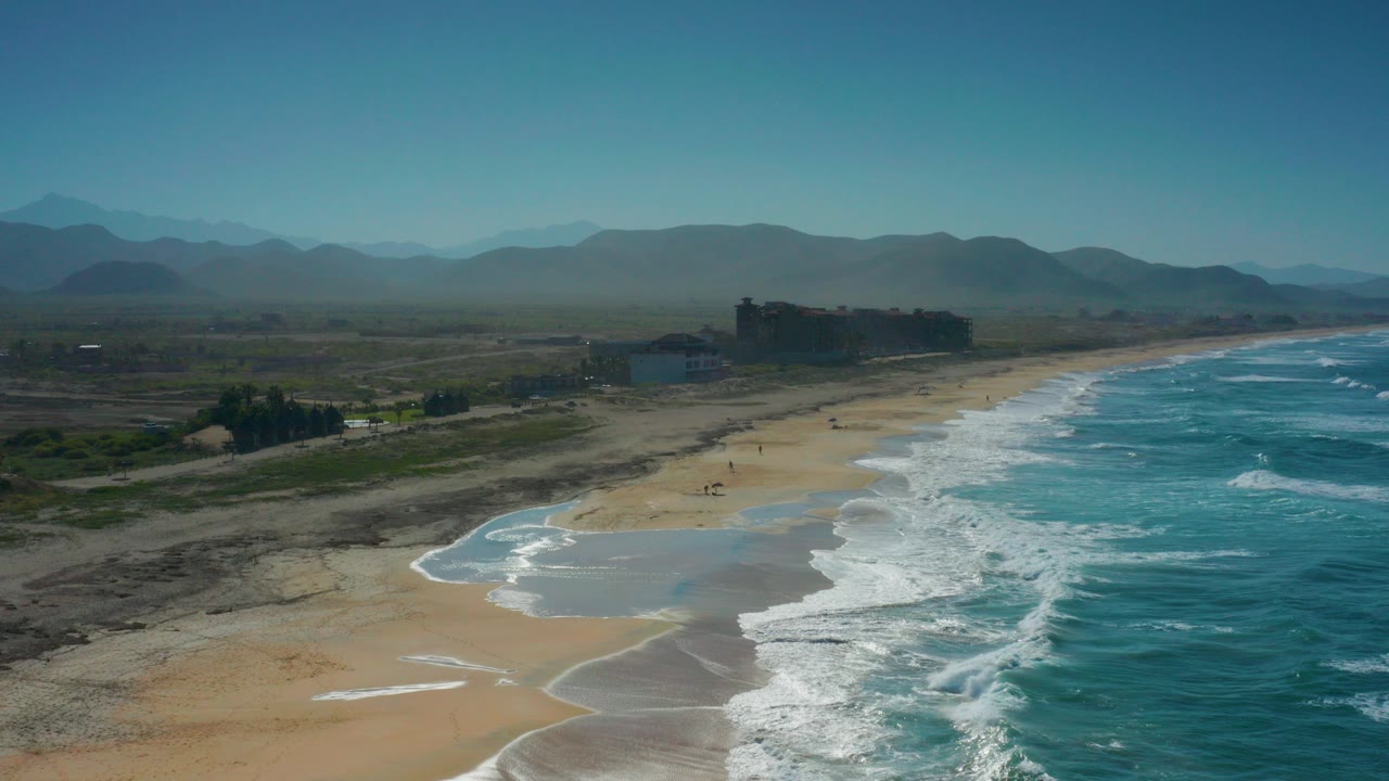 Aerial view over Cerritos, Baja California Sur with turquoise surf on the right, a broad sand beach, inland scrub, low shore buildings, distant mountains, and no visible people.