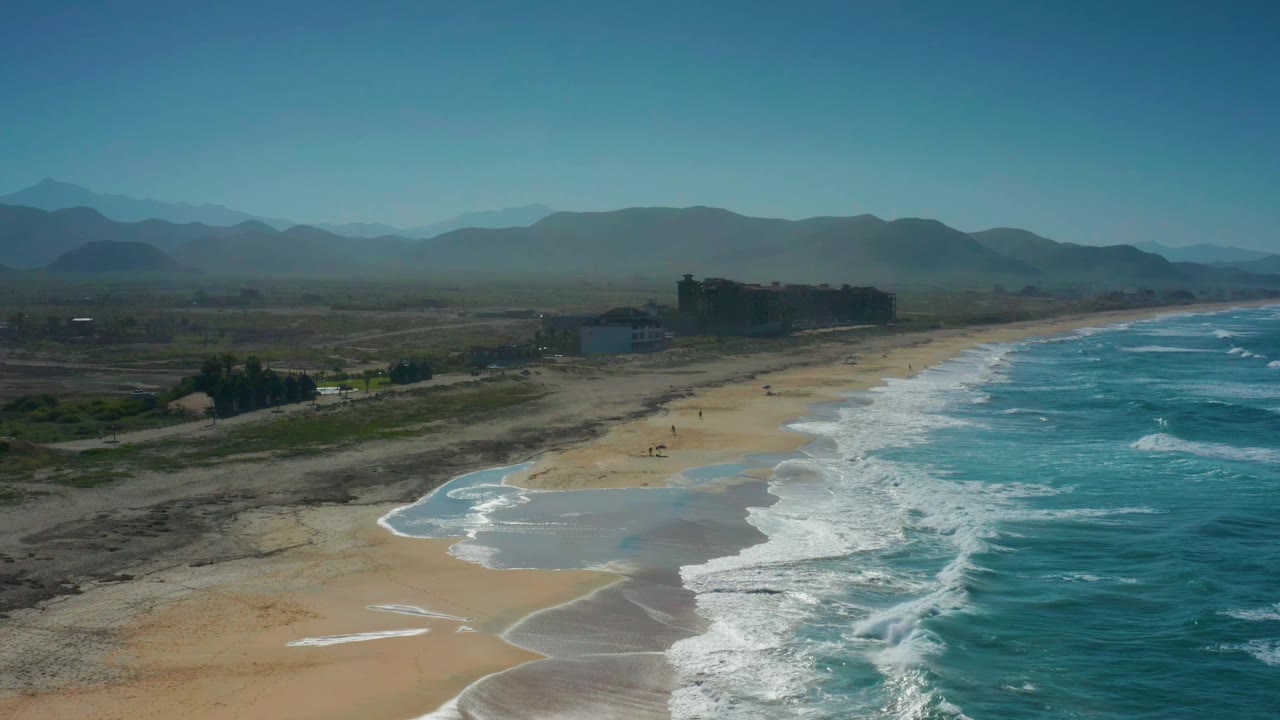 Wide aerial frame of Cerritos coastline with white waves breaking along the right edge, wet sand curves below, inland scrub to the left, shore buildings ahead, mountains in haze, and no visible people.