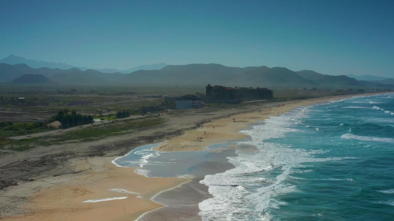 Aerial view in Cerritos showing brighter whitewater across the right side, pale sand and darker dune patches below, blocky buildings near the beach, hazy mountain layers, and no visible people.