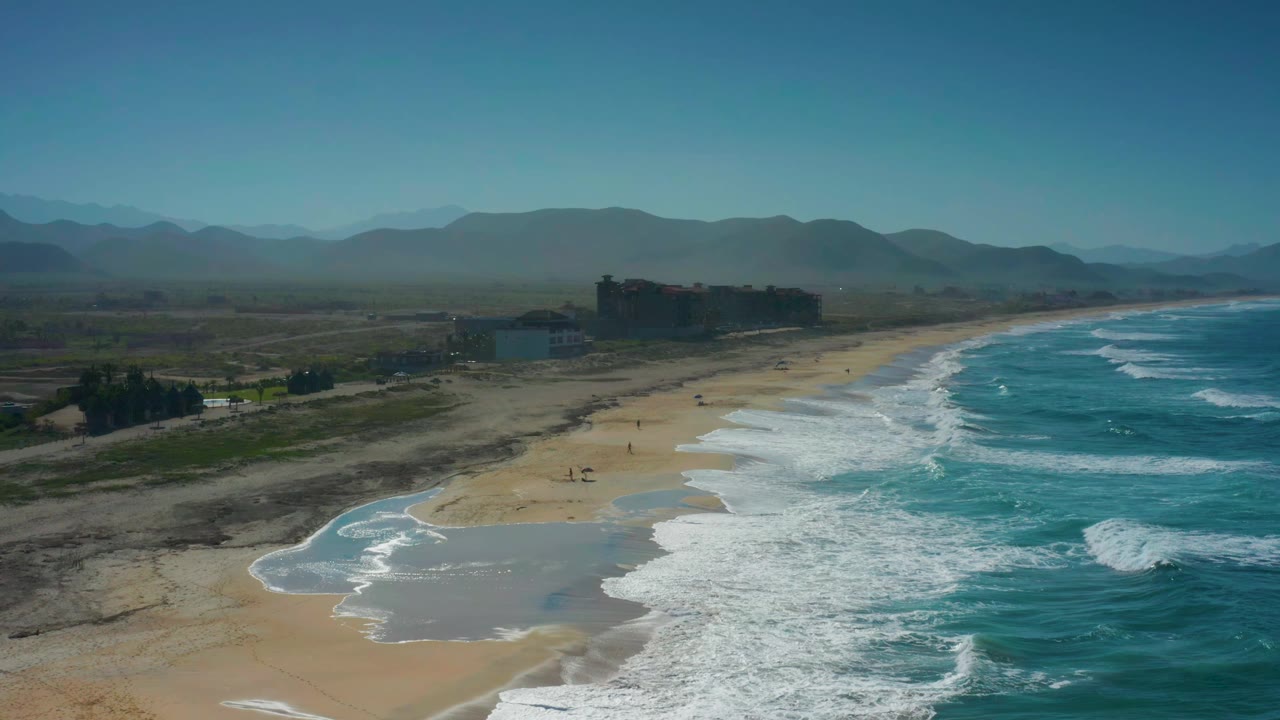 High aerial frame over Cerritos, Baja California Sur, showing white breakers edging the beach, shallow wet sand near the waterline, dark shoreline buildings, mountain ridges, and no visible people.
