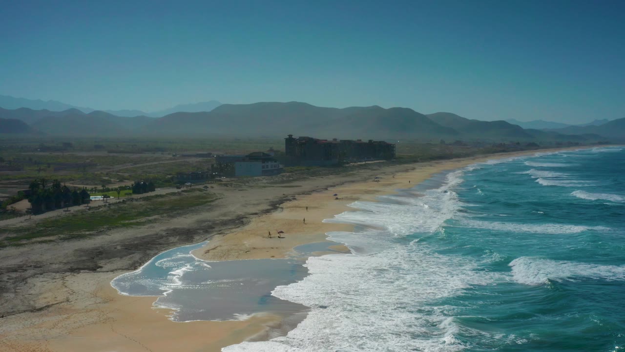 Aerial view along the Cerritos coastline with blue water filling the right side, foamy surf reaching the beach, sparse inland structures, hazy mountains, and no visible people.