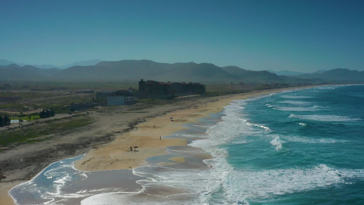 Aerial frame over Cerritos, Baja California Sur, showing blue-green surf meeting a broad sandy beach, low shore buildings, and hazy mountains with no clearly visible people.