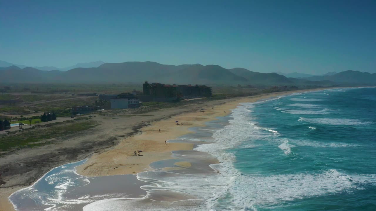 Wide drone view of a curving beach with white surf and distant mountains.