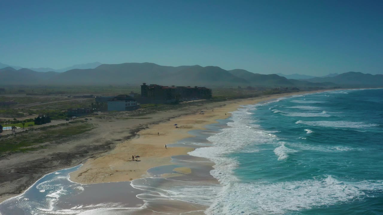 The aerial composition holds on Cerritos, with stronger white water in the lower right, open sand through the center, and low structures set back from the beach under the mountain line.