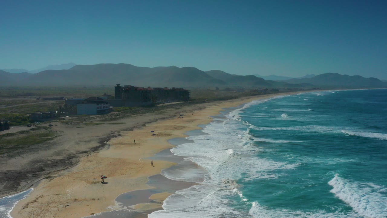 Aerial frame over Cerritos, Baja California Sur, showing white surf on a wide sandy beach, small beachgoers near the water, coastal buildings at left, and hazy mountains beyond.