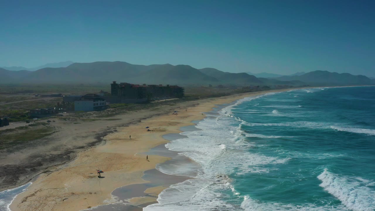 High coastal view of waves reaching a long beach beside low buildings.
