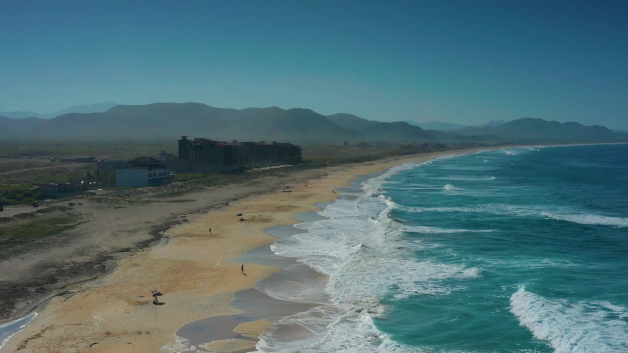 Cerritos, Baja California Sur beach seen from above, with bright blue water on the right, foam bands crossing the surf zone, several tiny people on the sand, and dark coastal buildings at left.