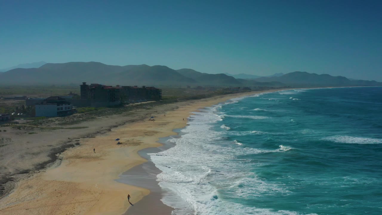 Wide aerial view of Cerritos, Baja California Sur, with white breakers pushing toward the sand, a darker building cluster inland left, mountains behind it, and tiny beachgoers visible below.