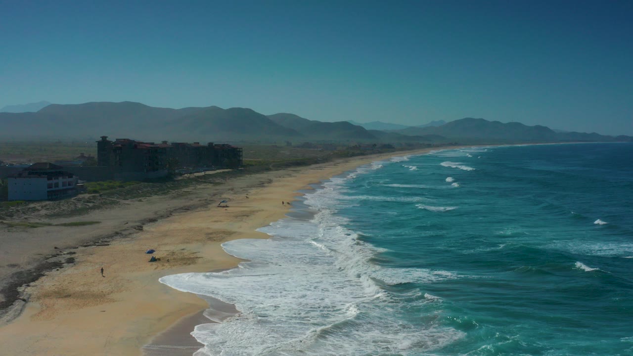 Wide aerial view over Cerritos, Baja California Sur, with turquoise waves curling onto a long sandy beach, low buildings near the dunes, hazy mountains beyond, and no visible people.