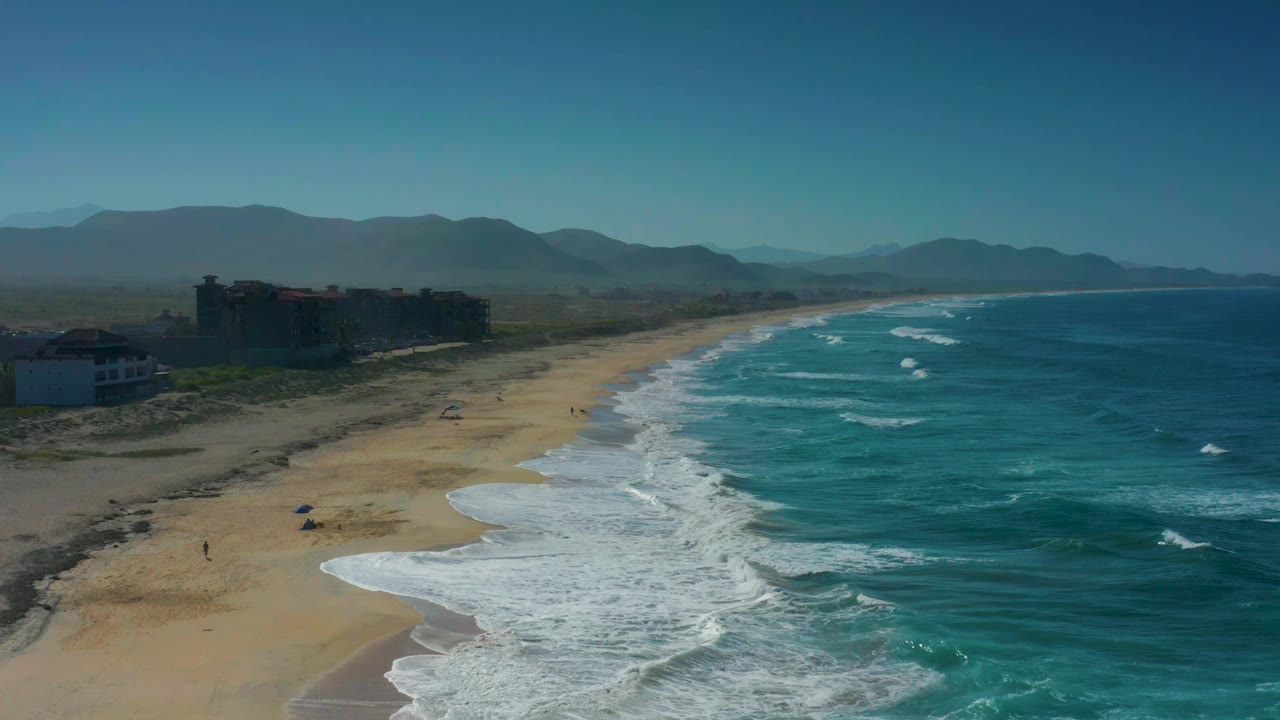 Aerial frame over Cerritos showing white wave lines advancing toward the beach, a cluster of low beachfront buildings at left, distant mountains on the horizon, and no visible people.