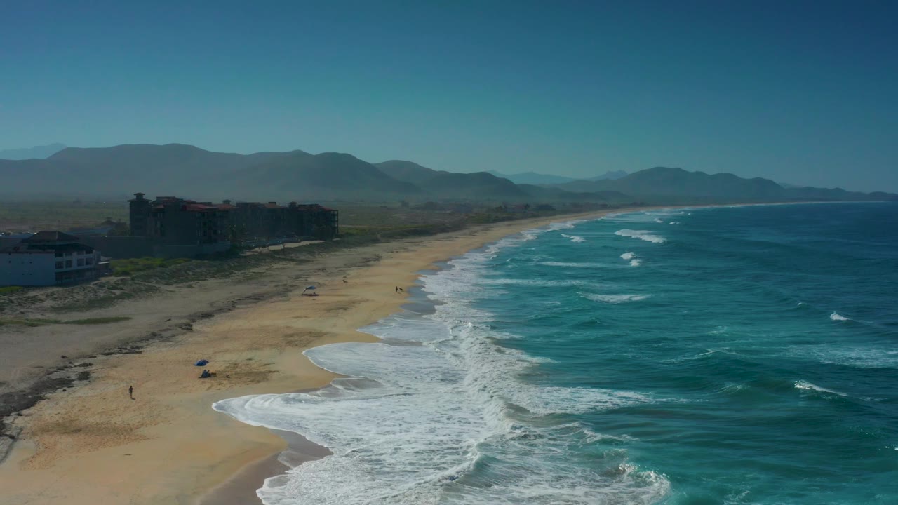 Elevated view of Cerritos coastline with the sandy beach curving into the distance, blue-green water on the right, foamy surf bands at midframe, and no visible people.