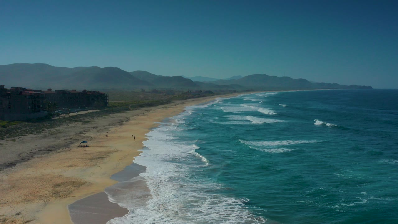 Aerial frame over Cerritos, Baja California Sur showing the sandy beach curving beside teal surf, dark buildings on the left edge, hazy mountains in the distance, and no visible people.