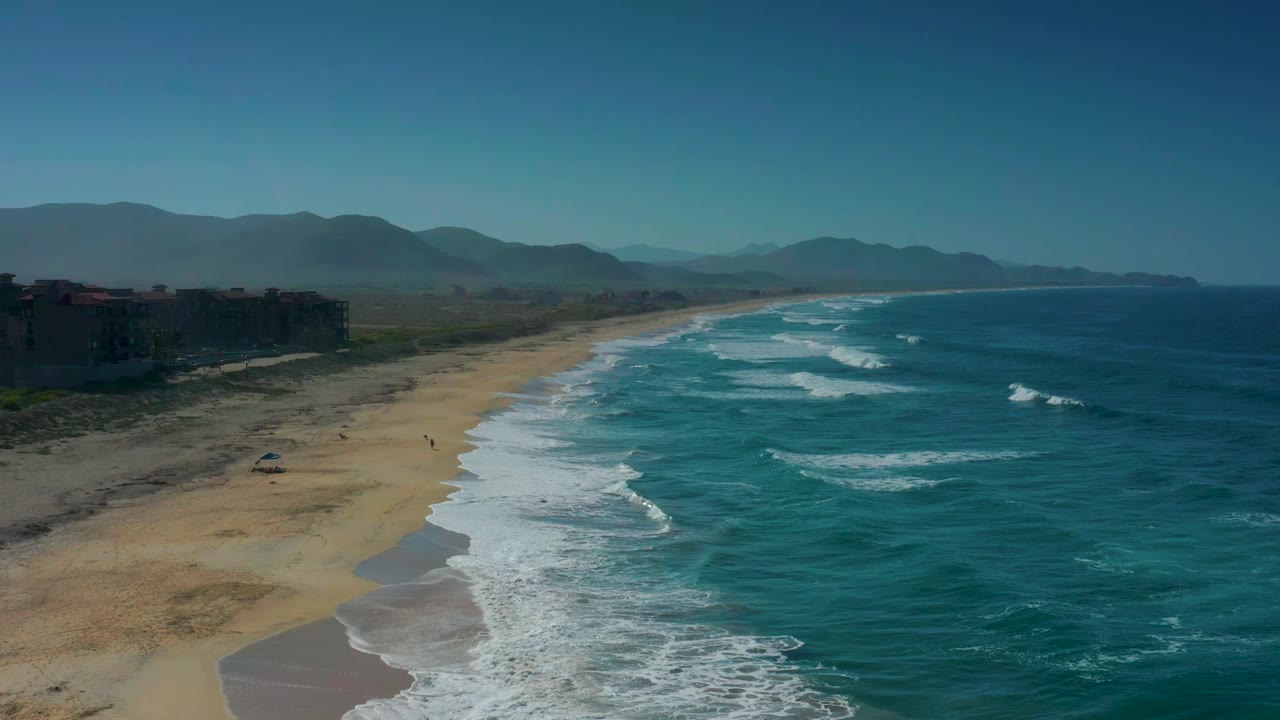 Cerritos beach from above with a longer sweep of white wave foam along the shore, ocean filling the right side, buildings at far left, mountains behind, and no visible people.